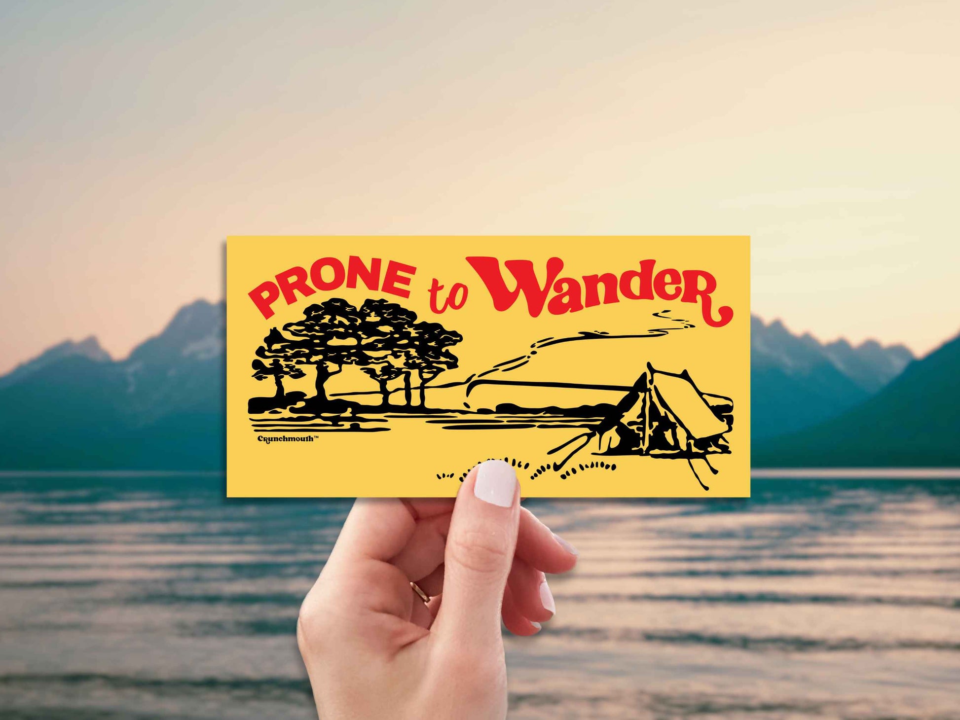 prone to wander camping bumper sticker, hand display, scenic mountains lake background