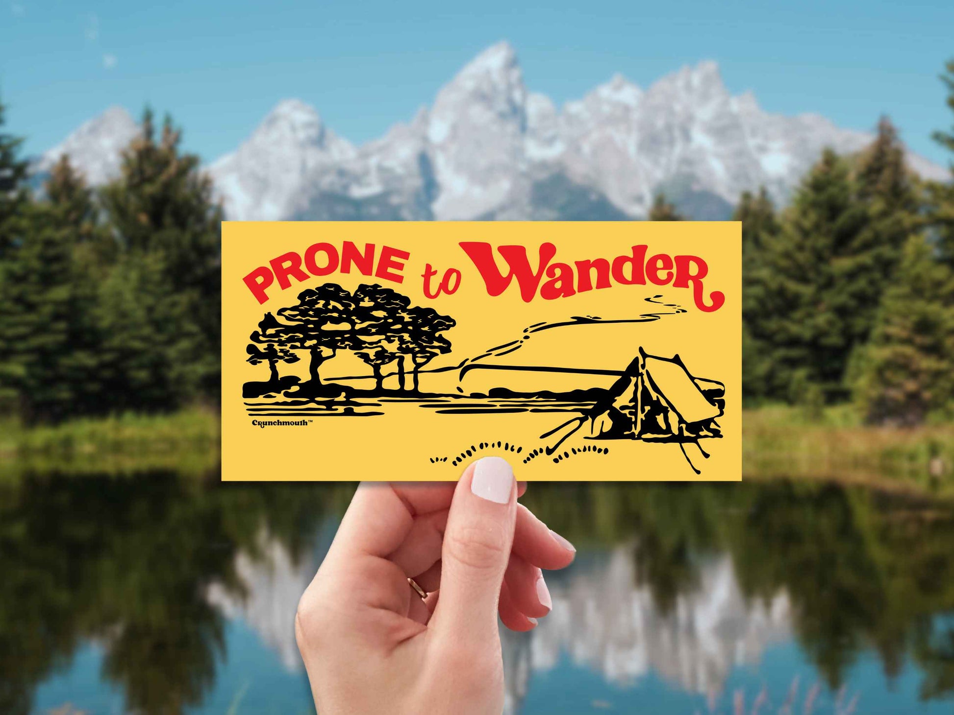 prone to wander camping bumper sticker, hand display, scenic mountains background