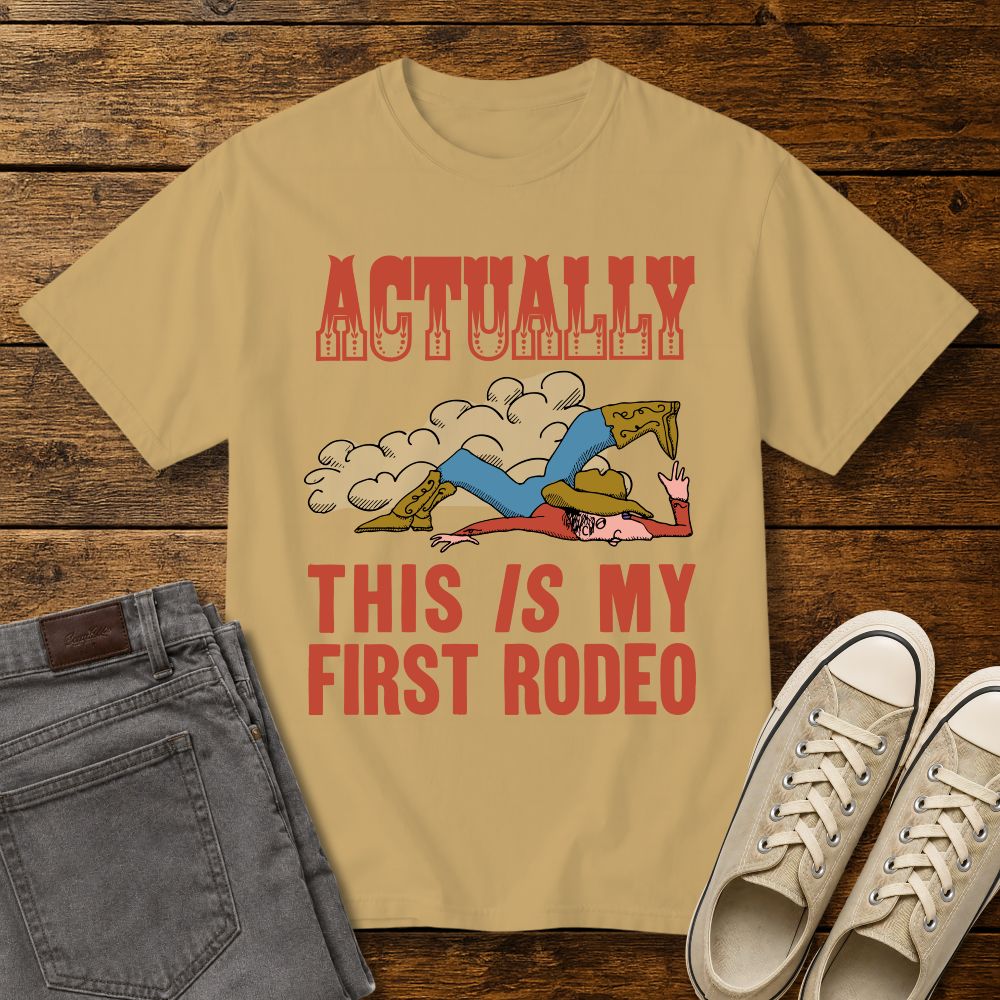 mustard graphic T-shirt mockup featuring a vintage-style cowboy illustration falling in the dirt with the phrase “Actually This Is My First Rodeo” in western-inspired red lettering, styled with gray jeans and beige sneakers on a rustic wood background.