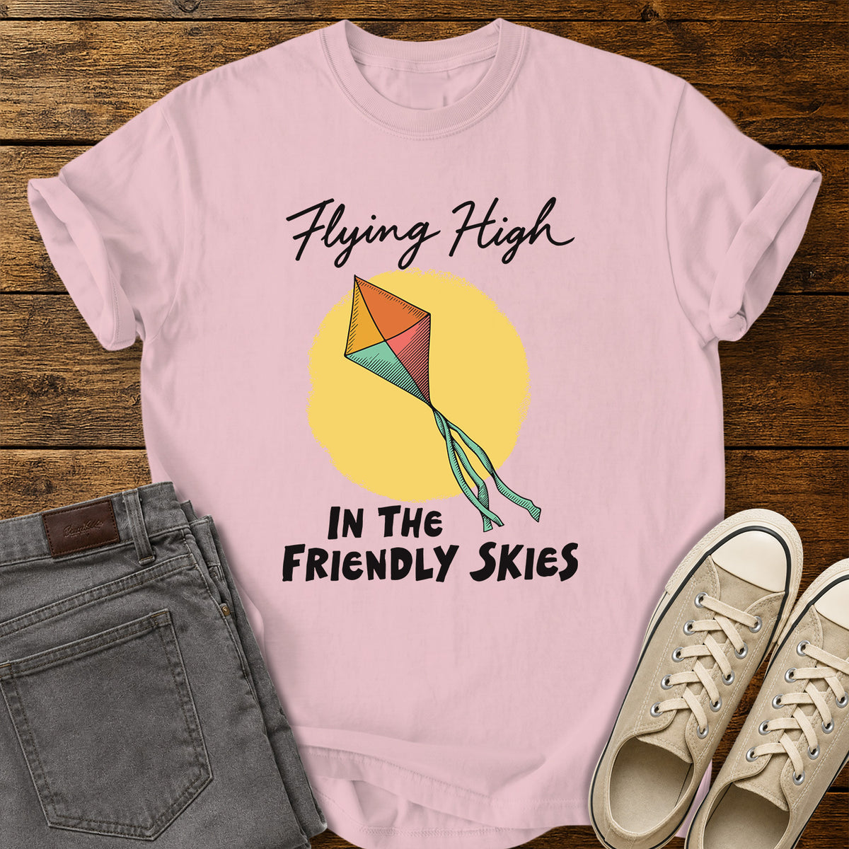 blossom pink T-shirt featuring a colorful retro kite flying against a yellow sun, with black script and bold lettering that reads “Flying High in the Friendly Skies,” displayed on a rustic wooden background with gray jeans and beige sneakers.