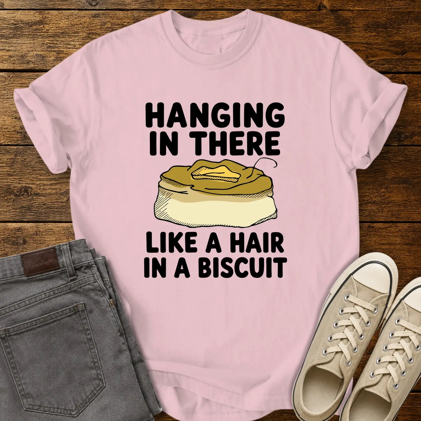 blossom pink T-shirt mockup featuring the phrase “HANGING IN THERE LIKE A HAIR IN A BISCUIT” in bold black lettering with a hand-drawn biscuit illustration and a single hair resting on top. The shirt is displayed on a rustic wood background alongside folded gray jeans and cream-colored canvas sneakers.