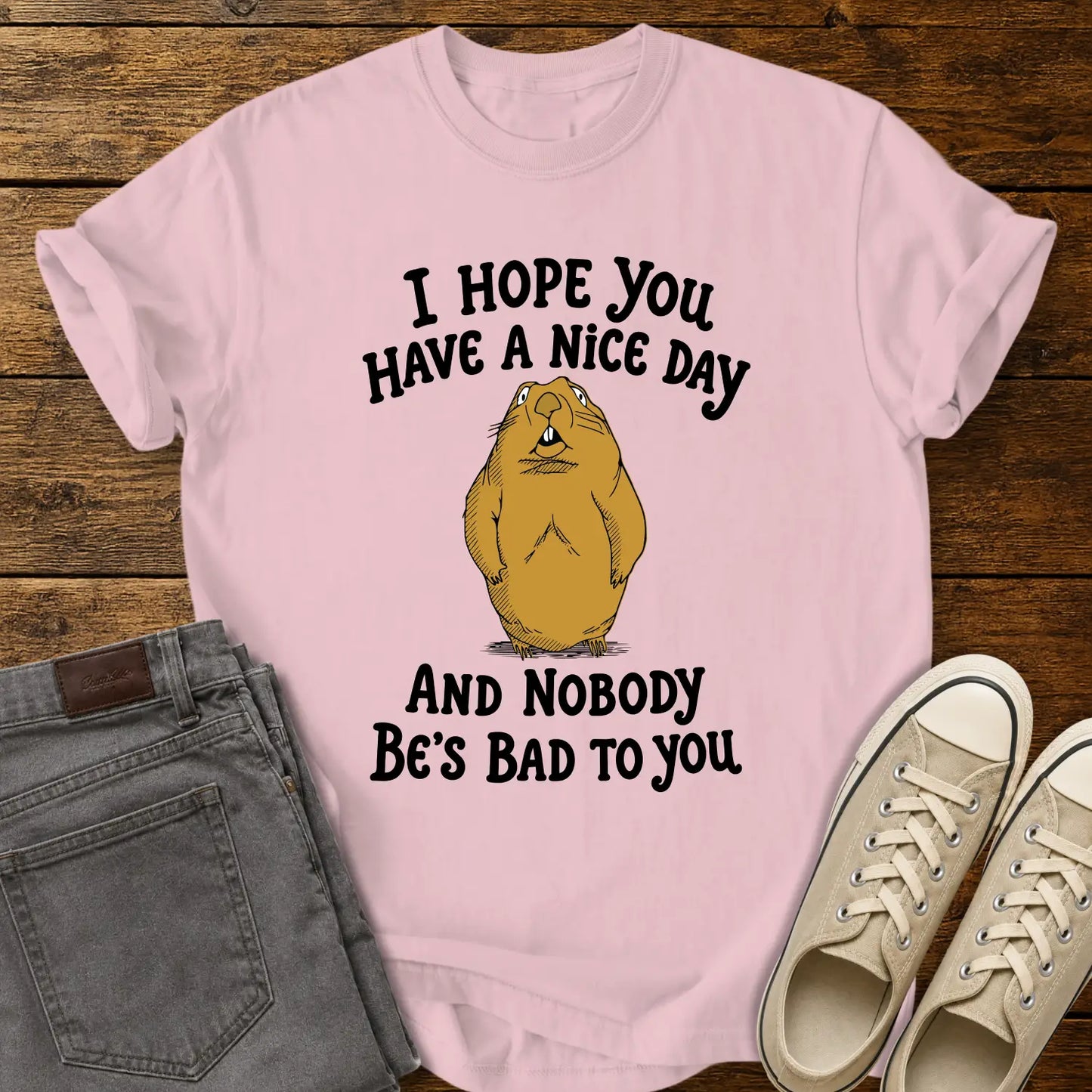 blossom pink T-shirt featuring a cartoon groundhog beneath the phrase “I HOPE YOU HAVE A NICE DAY AND NOBODY BE'S BAD TO YOU.” The shirt is displayed in a flat-lay on a rustic wood plank background with folded gray jeans and cream canvas sneakers.