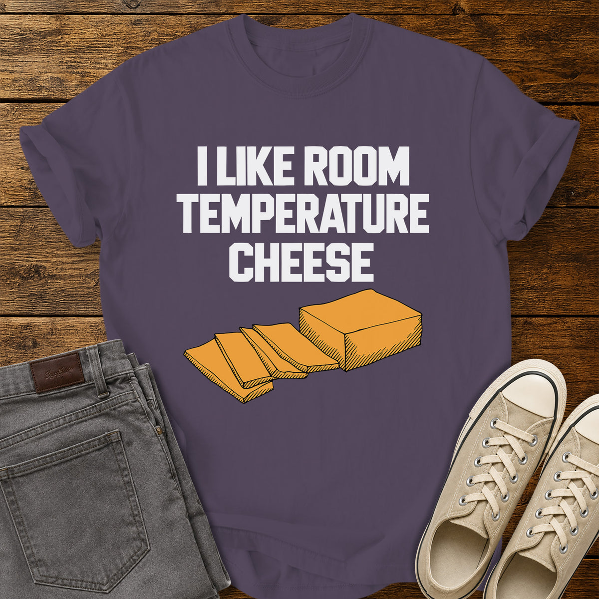 wine purple graphic T-shirt featuring bold white lettering reading “I Like Room Temperature Cheese” above an orange illustration of a block of cheese with several slices, displayed on a rustic wooden background with gray jeans and beige sneakers.