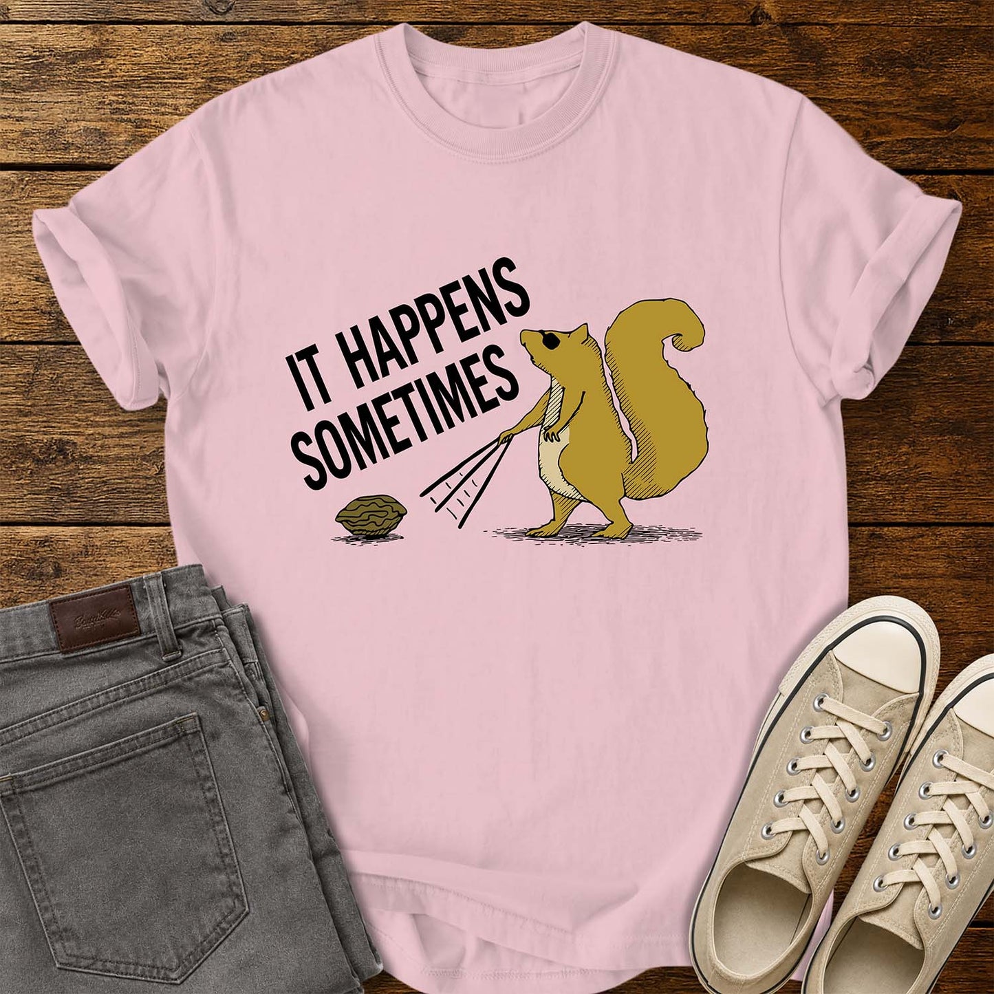 blossom pink T-shirt featuring a humorous retro illustration of a squirrel using a rake to pull a walnut toward itself. Bold black text angled above the artwork reads, “IT HAPPENS SOMETIMES.” The shirt is displayed on a rustic wood plank background with folded gray jeans on the left and cream canvas sneakers on the right.