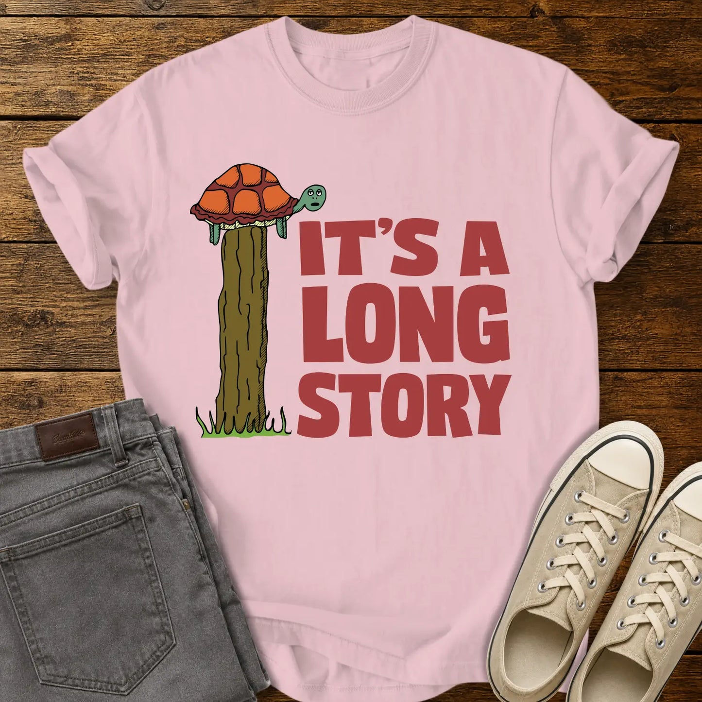 blossom pink T-shirt mockup featuring a humorous illustration of a turtle standing atop a fence post beside the phrase “IT’S A LONG STORY” in large vintage red lettering. The shirt is displayed on a rustic wood background alongside folded gray jeans and cream-colored canvas sneakers.
