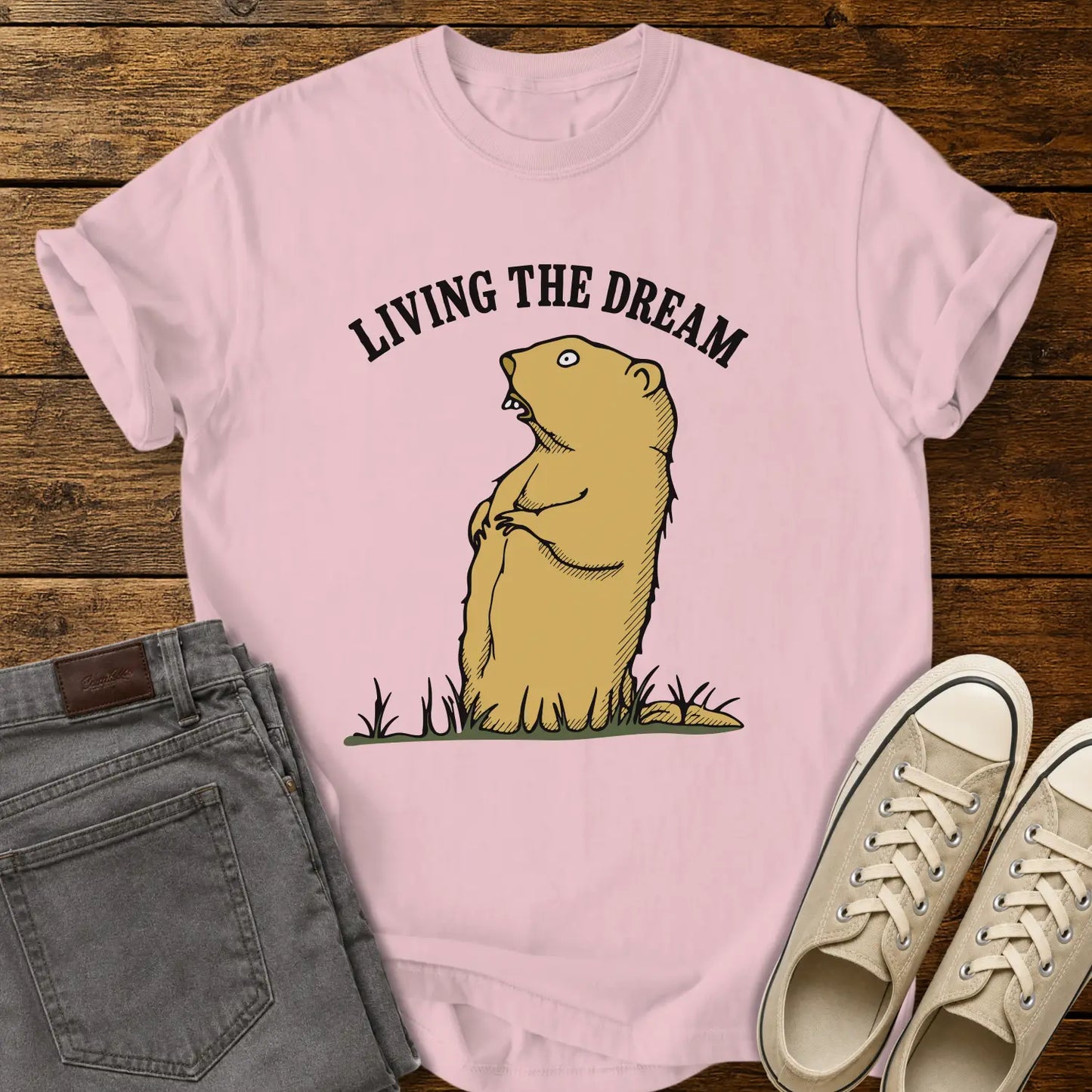 blossom pink graphic T-shirt mockup featuring a standing groundhog illustration beneath the phrase “Living the Dream” in bold black arched lettering, styled with gray jeans and beige sneakers on a rustic wood background.