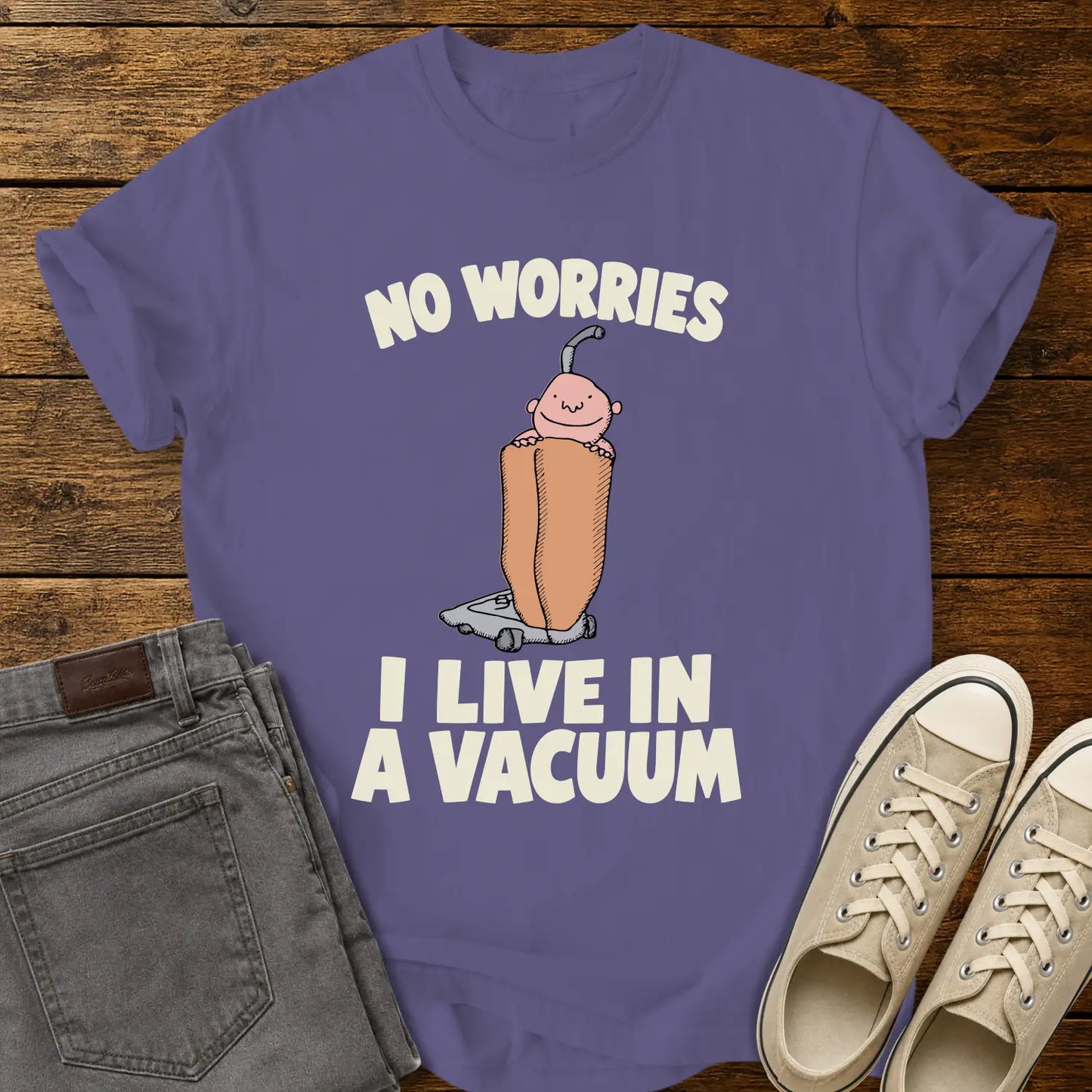 grape purple graphic T-shirt mockup featuring a cartoon person riding inside a vacuum cleaner bag with the phrase “No Worries, I Live in a Vacuum” in bold white lettering, styled with gray jeans and beige sneakers on a rustic wood background.