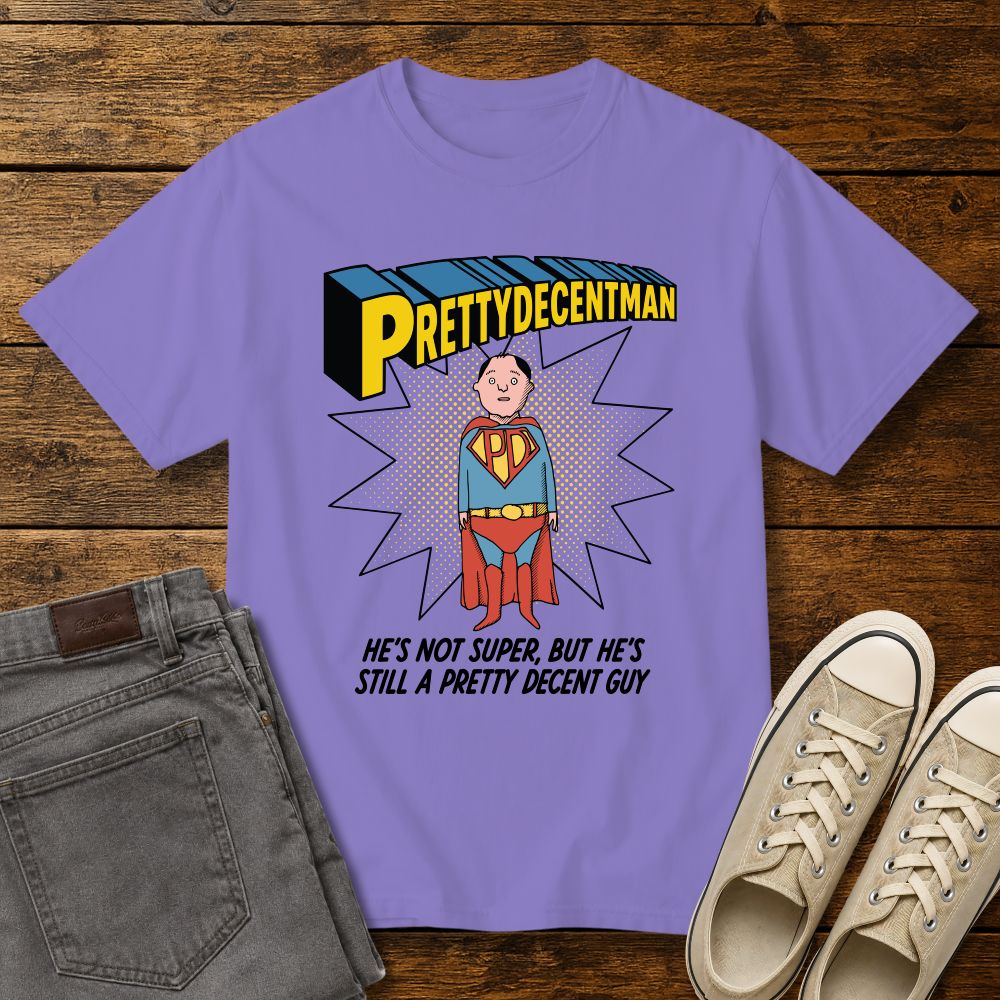 violet t-shirt mockup featuring a comic-style “PRETTYDECENTMAN” superhero graphic with a hand-drawn character in a blue and red costume standing in front of a halftone starburst. Text below reads: “HE’S NOT SUPER, BUT HE’S STILL A PRETTY DECENT GUY.” Shirt is displayed on a rustic wood background with gray jeans and beige sneakers.