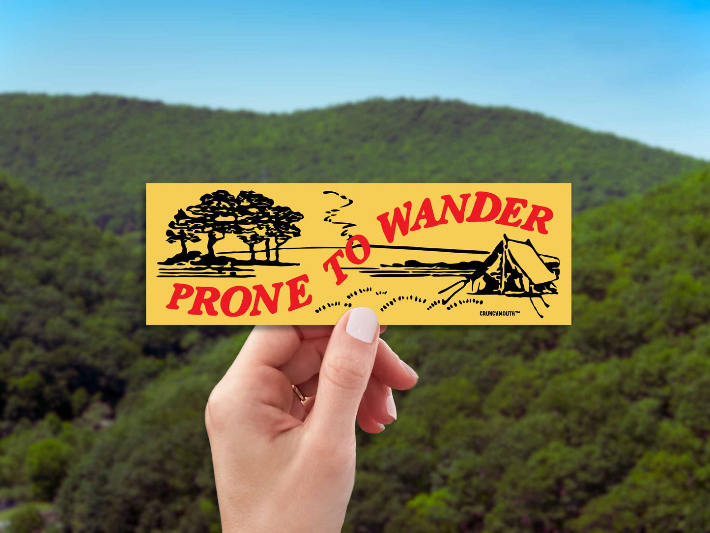 prone to wander camping bumper sticker, hand display, green mountains background