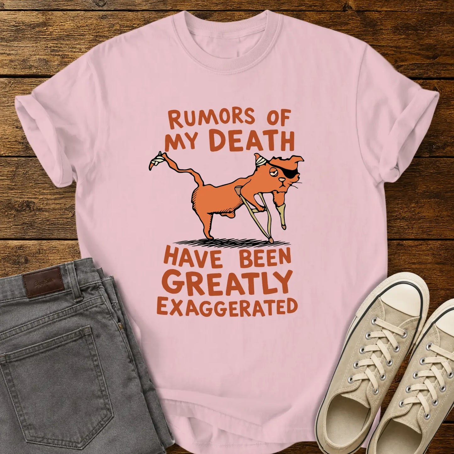 blossom pink T-shirt mockup featuring a humorous illustration of an orange cat with a peg leg, eye patch, and bandages alongside the phrase “RUMORS OF MY DEATH HAVE BEEN GREATLY EXAGGERATED” in retro orange lettering. The shirt is displayed on a rustic wood background alongside folded gray jeans and cream-colored canvas sneakers.