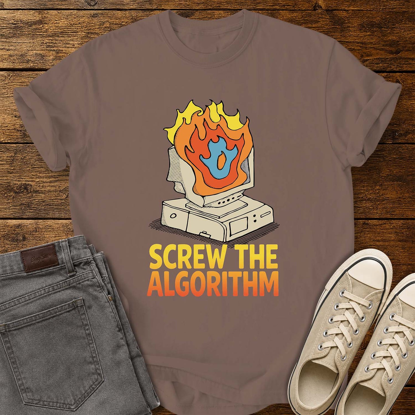 espresso brown T-shirt featuring a retro illustration of a vintage desktop computer with flames erupting from the monitor and a blue flame icon at the center. Beneath the graphic, bold yellow and orange text reads “SCREW THE ALGORITHM.” The shirt is displayed on a rustic wood plank background with folded gray jeans and a pair of cream canvas sneakers beside it.