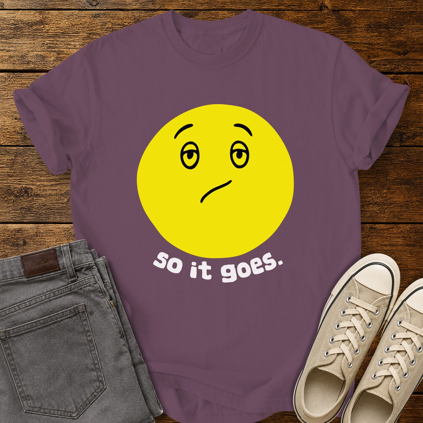 berry purple flat-lay t-shirt mockup featuring a large yellow retro-style smiley face with a subtly uncertain expression above the phrase, "so it goes." in rounded white lowercase lettering. The minimalist vintage-inspired design is centered on the shirt, which is displayed on a rustic wood plank background alongside folded gray jeans and a pair of cream canvas sneakers.