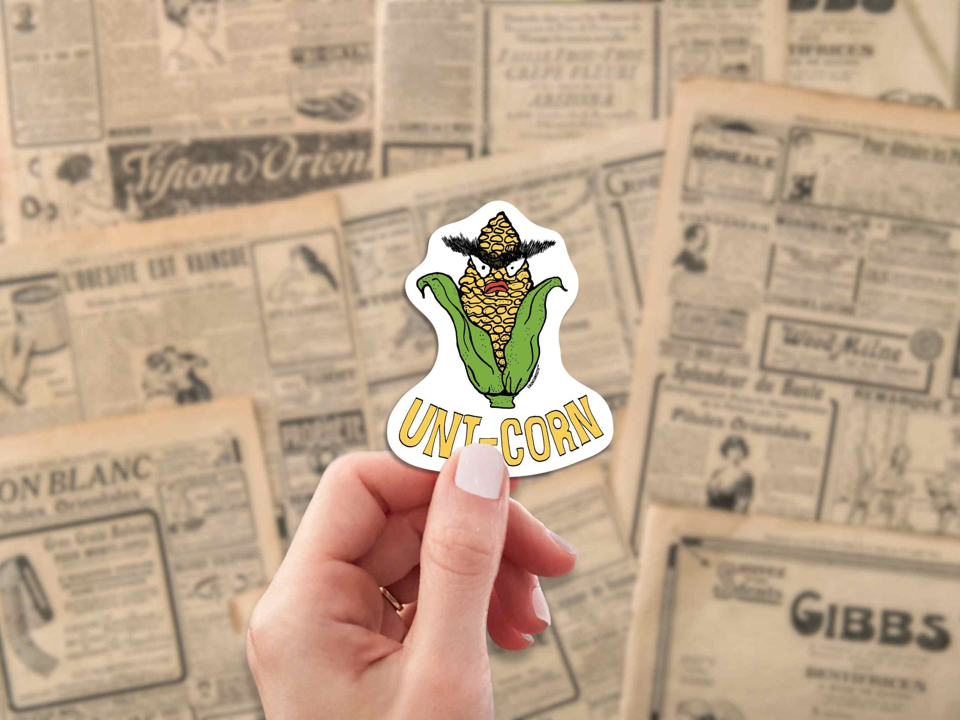uni-corn laptop sticker, ear of corn with unibrow, hand display, antique newspaper ads background