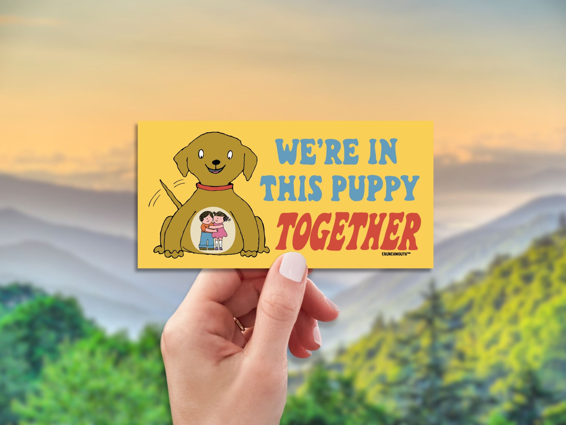 we're in this puppy together bumper sticker, hand display, blurred landscape background