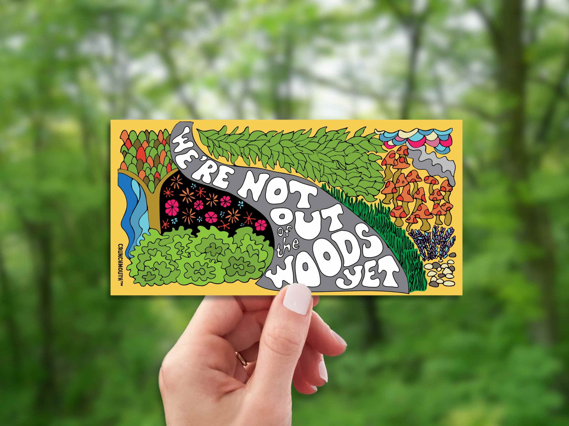 we're not out of the woods yet bumper sticker, hand display, blurred landscape background