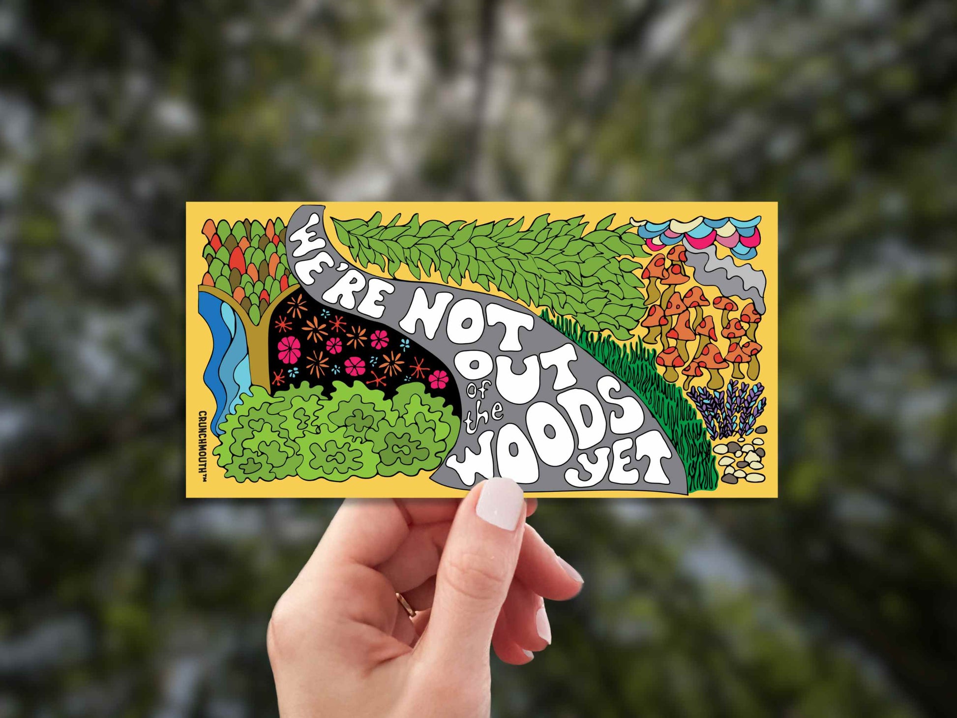 we're not out of the woods yet bumper sticker, hand display, blurred landscape background