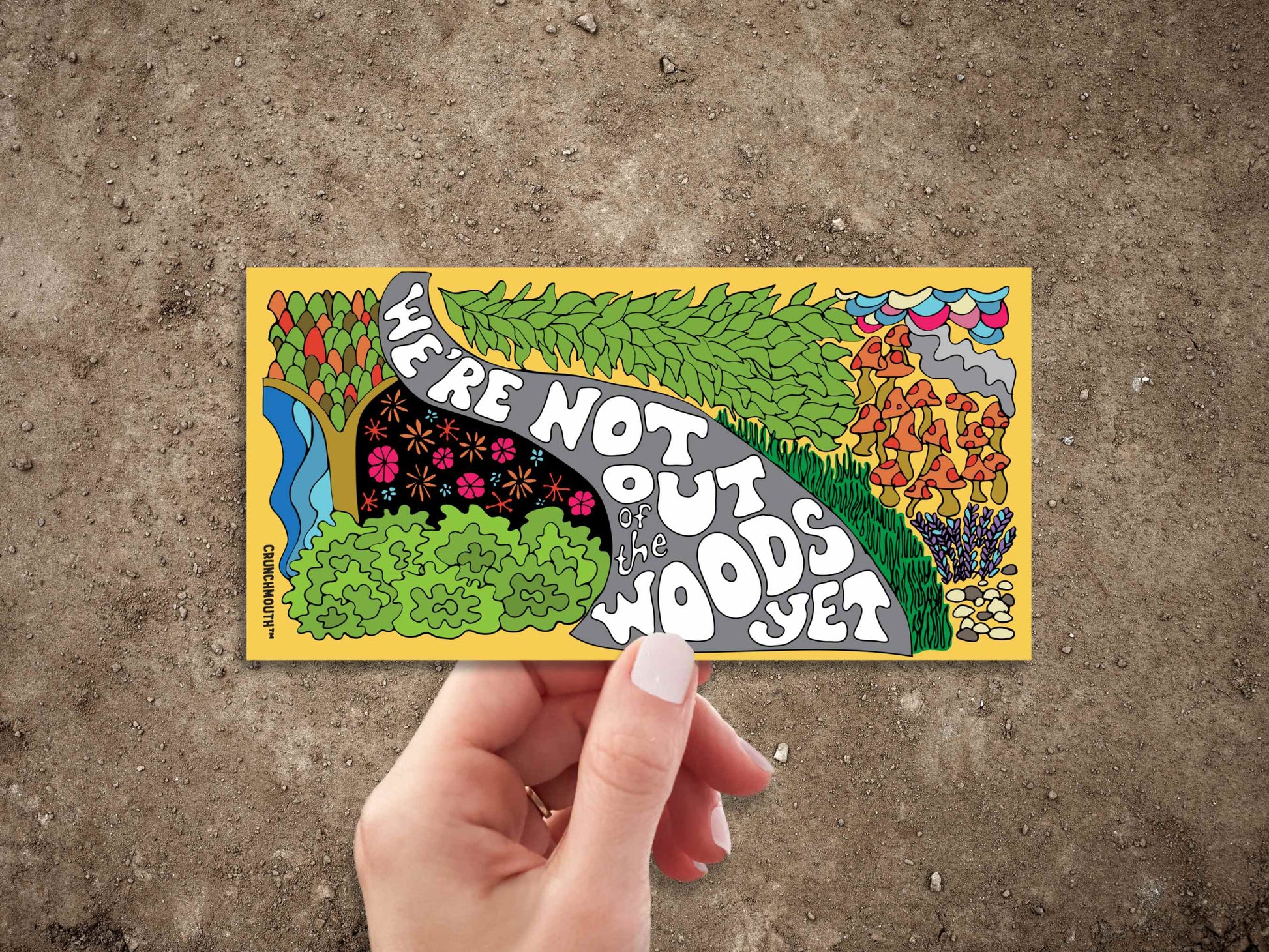 we're not out of the woods yet bumper sticker, hand display, dirt background