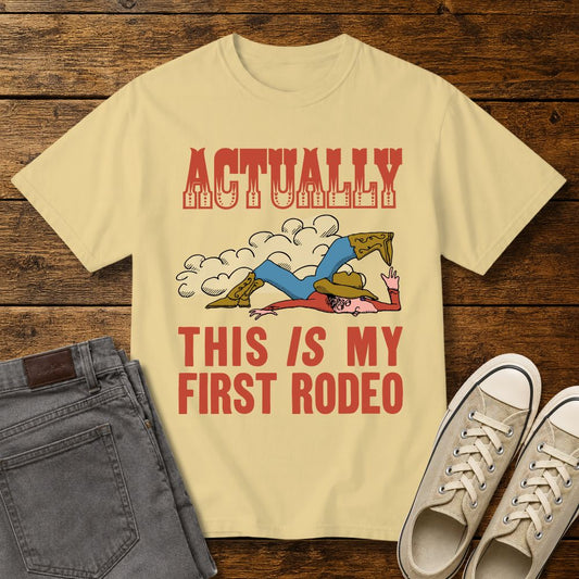 Butter yellow graphic T-shirt mockup featuring a vintage-style cowboy illustration falling in the dirt with the phrase “Actually This Is My First Rodeo” in western-inspired red lettering, styled with gray jeans and beige sneakers on a rustic wood background.