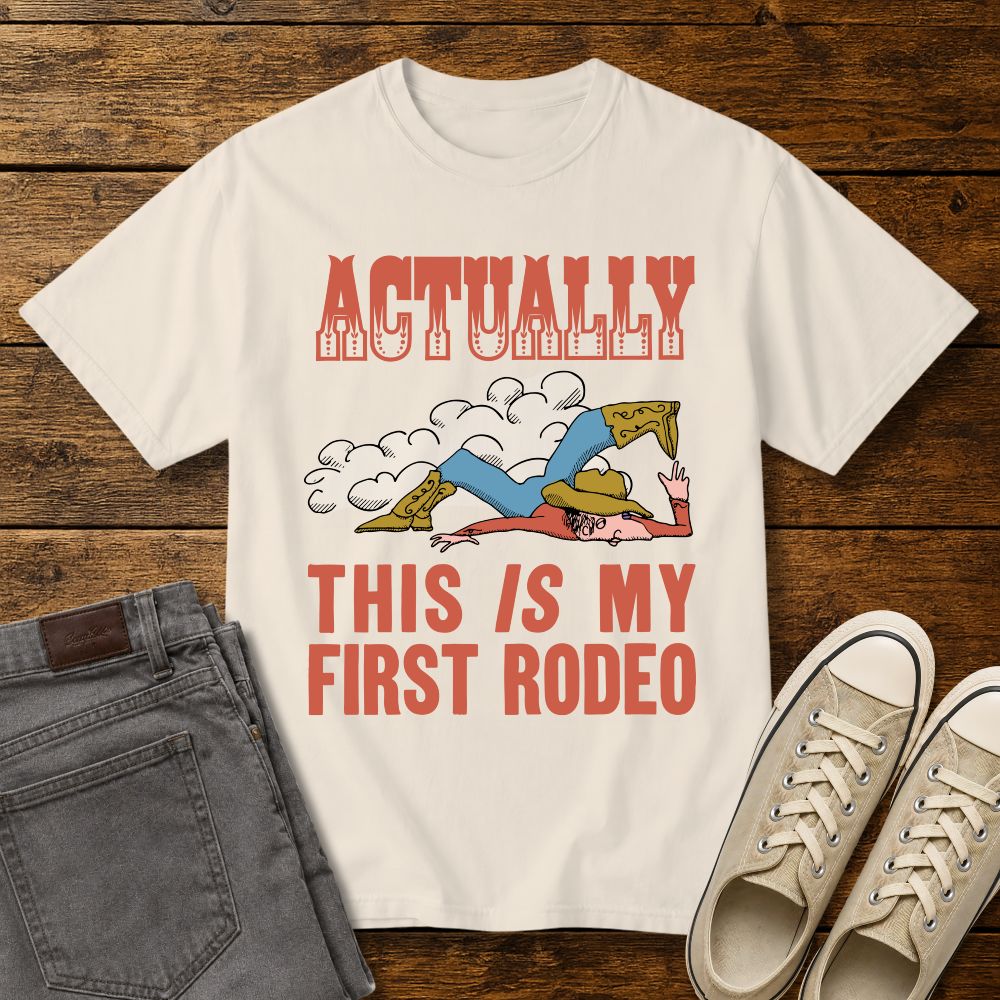 ivory graphic T-shirt mockup featuring a vintage-style cowboy illustration falling in the dirt with the phrase “Actually This Is My First Rodeo” in western-inspired red lettering, styled with gray jeans and beige sneakers on a rustic wood background.