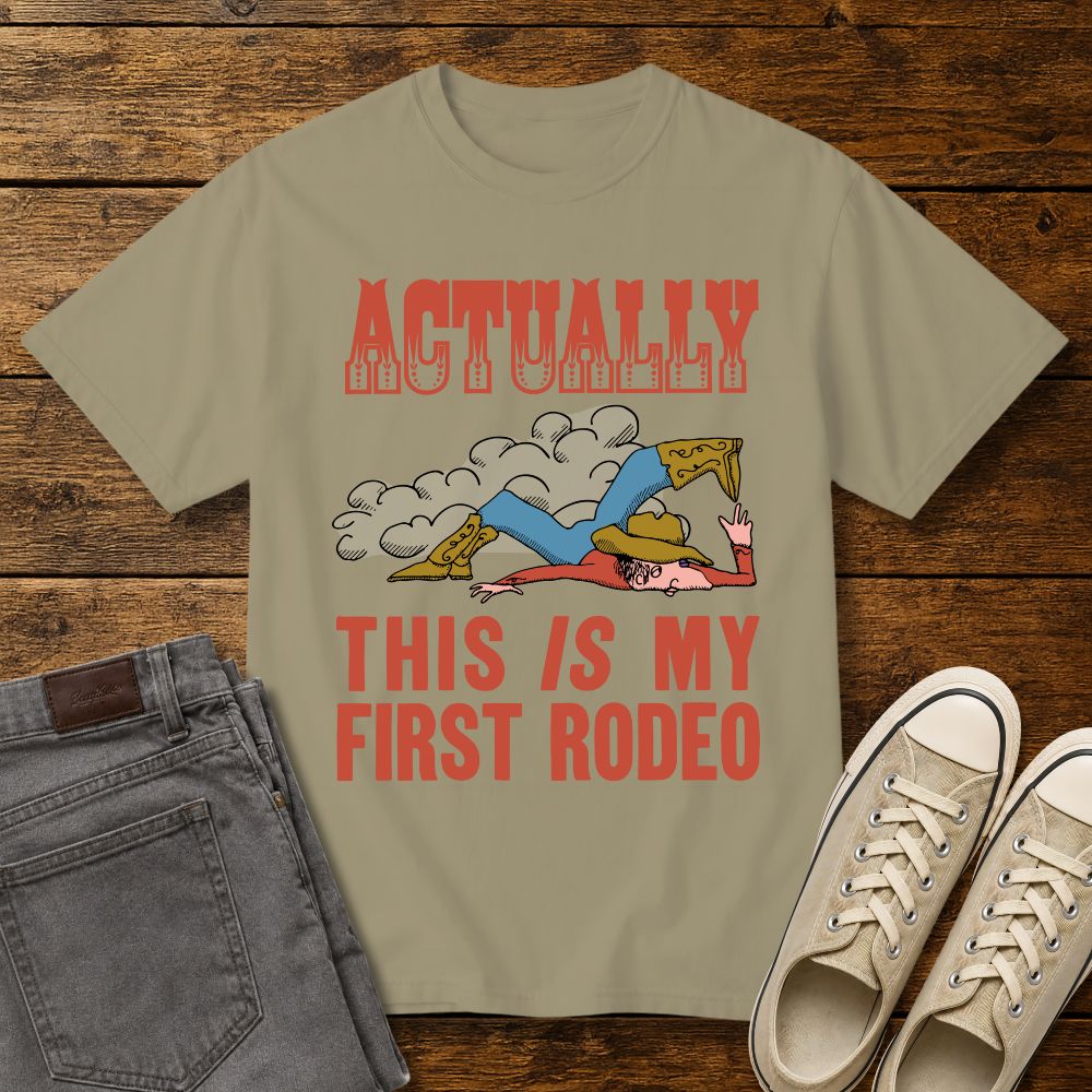 khaki graphic T-shirt mockup featuring a vintage-style cowboy illustration falling in the dirt with the phrase “Actually This Is My First Rodeo” in western-inspired red lettering, styled with gray jeans and beige sneakers on a rustic wood background.