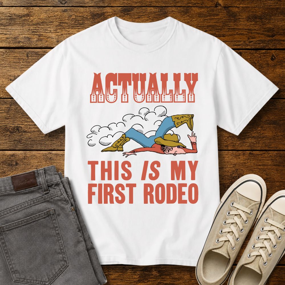 white graphic T-shirt mockup featuring a vintage-style cowboy illustration falling in the dirt with the phrase “Actually This Is My First Rodeo” in western-inspired red lettering, styled with gray jeans and beige sneakers on a rustic wood background.