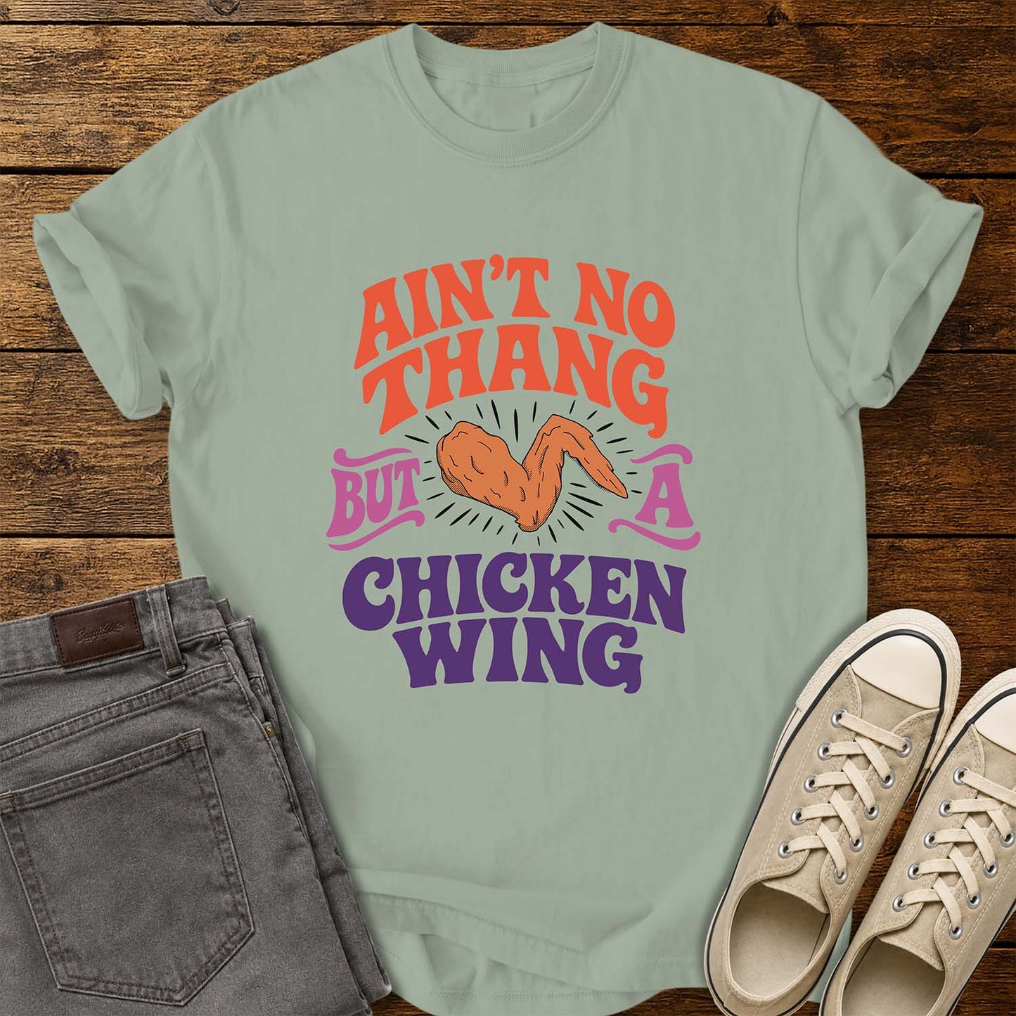 bay green flat-lay t-shirt mockup featuring colorful retro lettering that reads, "AIN'T NO THANG BUT A CHICKEN WING," with a cartoon chicken wing illustrated in the center and accented by radiating lines. The vintage-inspired design uses coral, purple, and pink lettering for a fun, nostalgic look. The shirt is displayed on a rustic wood plank background alongside folded gray jeans and a pair of cream canvas sneakers.