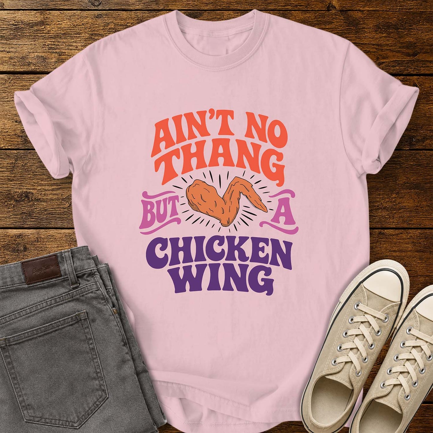 blossom pink flat-lay t-shirt mockup featuring colorful retro lettering that reads, "AIN'T NO THANG BUT A CHICKEN WING," with a cartoon chicken wing illustrated in the center and accented by radiating lines. The vintage-inspired design uses coral, purple, and pink lettering for a fun, nostalgic look. The shirt is displayed on a rustic wood plank background alongside folded gray jeans and a pair of cream canvas sneakers.
