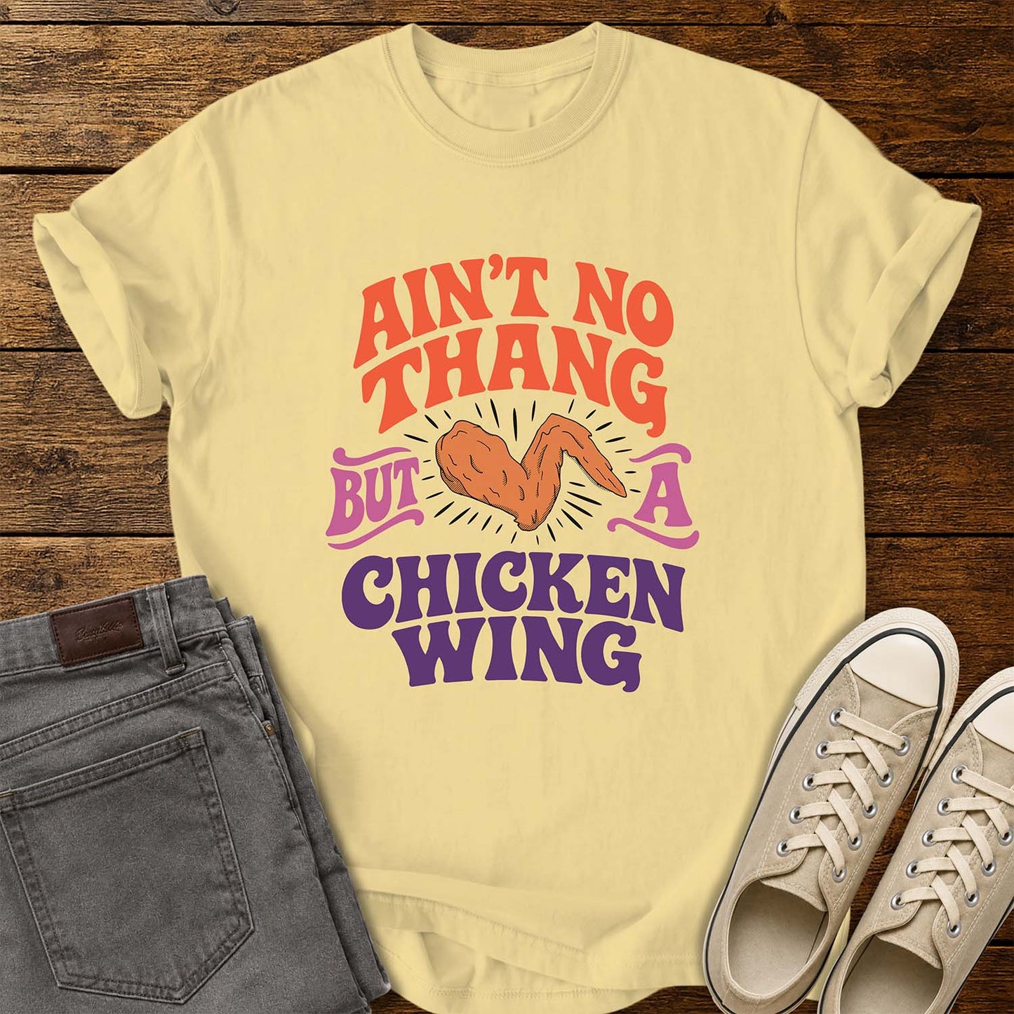 butter yellow flat-lay t-shirt mockup featuring colorful retro lettering that reads, "AIN'T NO THANG BUT A CHICKEN WING," with a cartoon chicken wing illustrated in the center and accented by radiating lines. The vintage-inspired design uses coral, purple, and pink lettering for a fun, nostalgic look. The shirt is displayed on a rustic wood plank background alongside folded gray jeans and a pair of cream canvas sneakers.