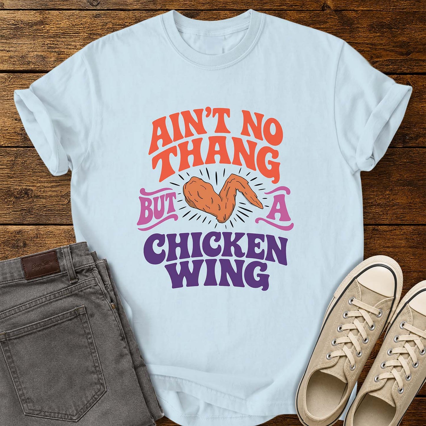 chambray blue flat-lay t-shirt mockup featuring colorful retro lettering that reads, "AIN'T NO THANG BUT A CHICKEN WING," with a cartoon chicken wing illustrated in the center and accented by radiating lines. The vintage-inspired design uses coral, purple, and pink lettering for a fun, nostalgic look. The shirt is displayed on a rustic wood plank background alongside folded gray jeans and a pair of cream canvas sneakers.