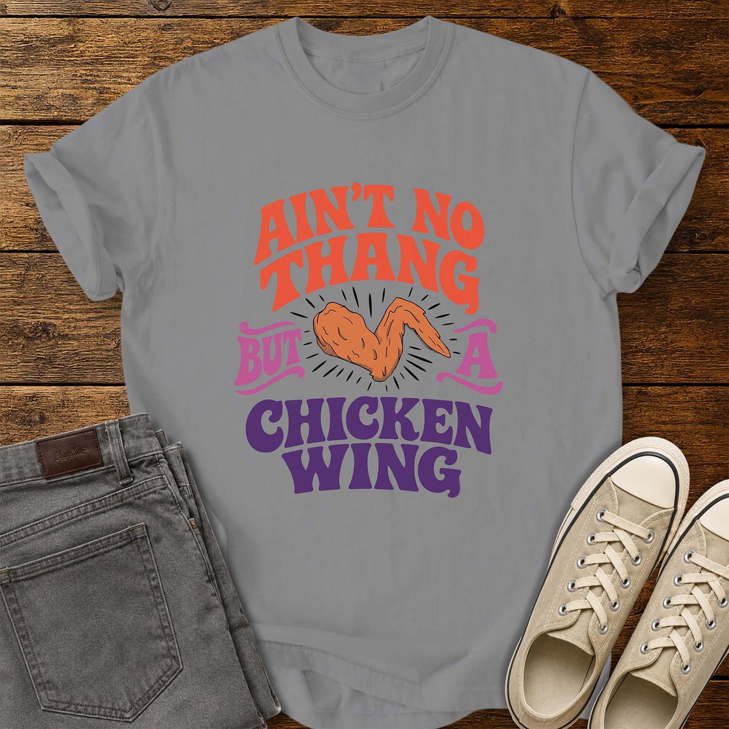 grey flat-lay t-shirt mockup featuring colorful retro lettering that reads, "AIN'T NO THANG BUT A CHICKEN WING," with a cartoon chicken wing illustrated in the center and accented by radiating lines. The vintage-inspired design uses coral, purple, and pink lettering for a fun, nostalgic look. The shirt is displayed on a rustic wood plank background alongside folded gray jeans and a pair of cream canvas sneakers.
