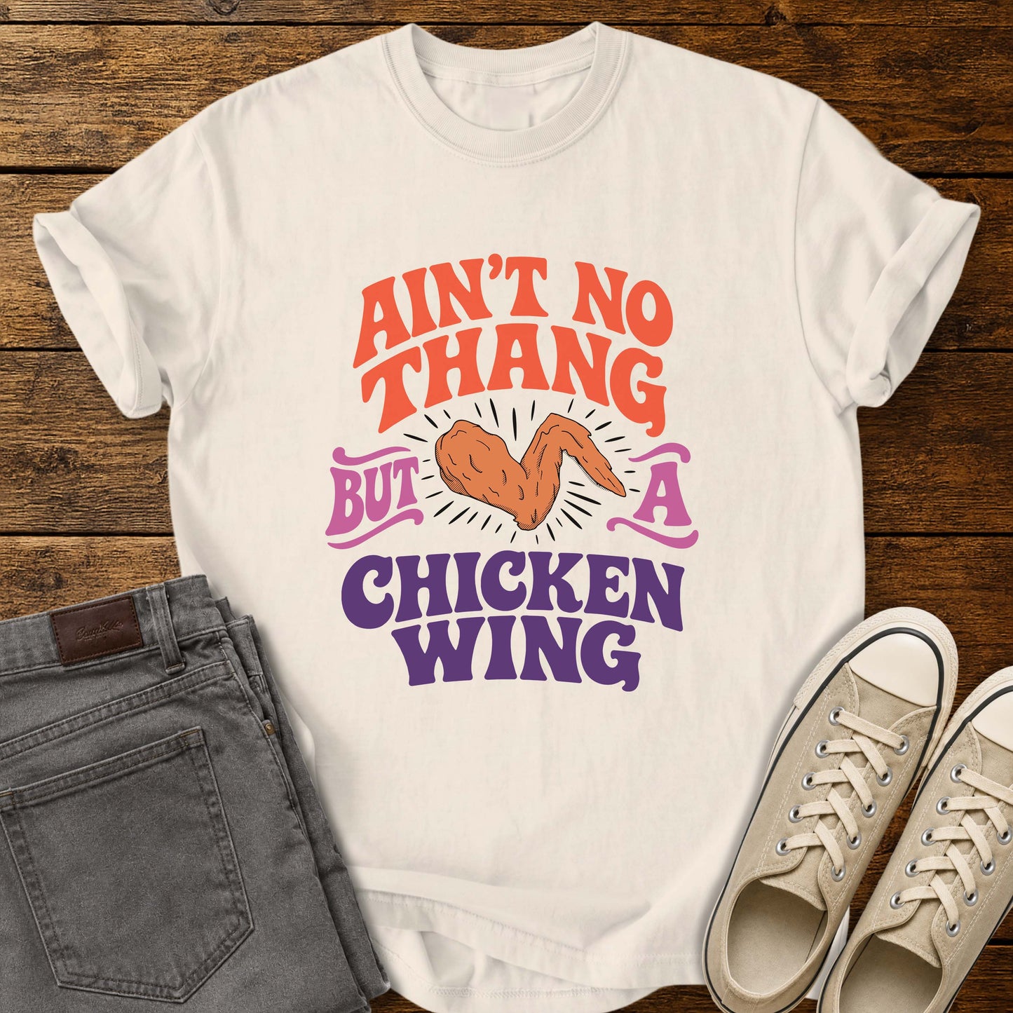 Ivory flat-lay t-shirt mockup featuring colorful retro lettering that reads, "AIN'T NO THANG BUT A CHICKEN WING," with a cartoon chicken wing illustrated in the center and accented by radiating lines. The vintage-inspired design uses coral, purple, and pink lettering for a fun, nostalgic look. The shirt is displayed on a rustic wood plank background alongside folded gray jeans and a pair of cream canvas sneakers.