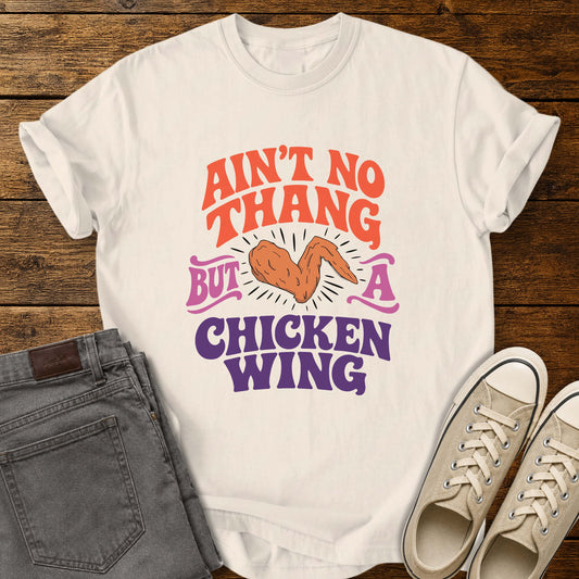 Ivory flat-lay t-shirt mockup featuring colorful retro lettering that reads, "AIN'T NO THANG BUT A CHICKEN WING," with a cartoon chicken wing illustrated in the center and accented by radiating lines. The vintage-inspired design uses coral, purple, and pink lettering for a fun, nostalgic look. The shirt is displayed on a rustic wood plank background alongside folded gray jeans and a pair of cream canvas sneakers.