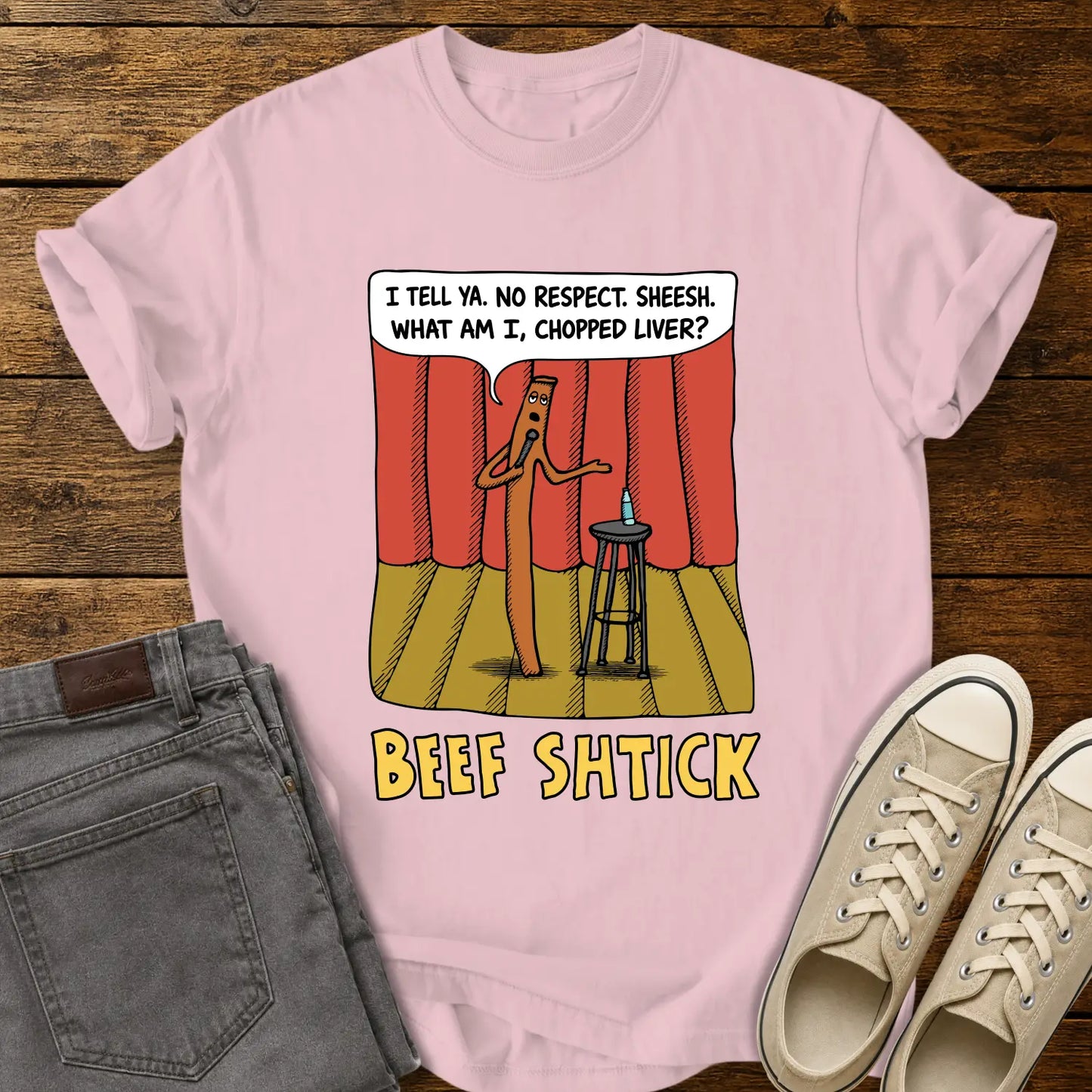 blossom pink T-shirt featuring a humorous cartoon of a beef stick performing stand-up comedy on a stage with red curtains, a stool, microphone, and water bottle. A speech bubble reads, “I tell ya. No respect. Sheesh. What am I, chopped liver?” The design is titled “BEEF SHTICK” in bold yellow lettering. The shirt is displayed on a rustic wood plank background with folded gray jeans and cream canvas sneakers.