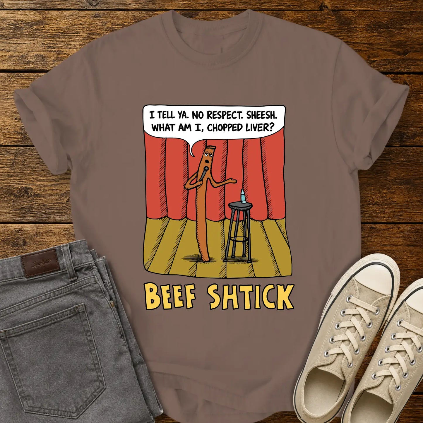 espresso T-shirt featuring a humorous cartoon of a beef stick performing stand-up comedy on a stage with red curtains, a stool, microphone, and water bottle. A speech bubble reads, “I tell ya. No respect. Sheesh. What am I, chopped liver?” The design is titled “BEEF SHTICK” in bold yellow lettering. The shirt is displayed on a rustic wood plank background with folded gray jeans and cream canvas sneakers.