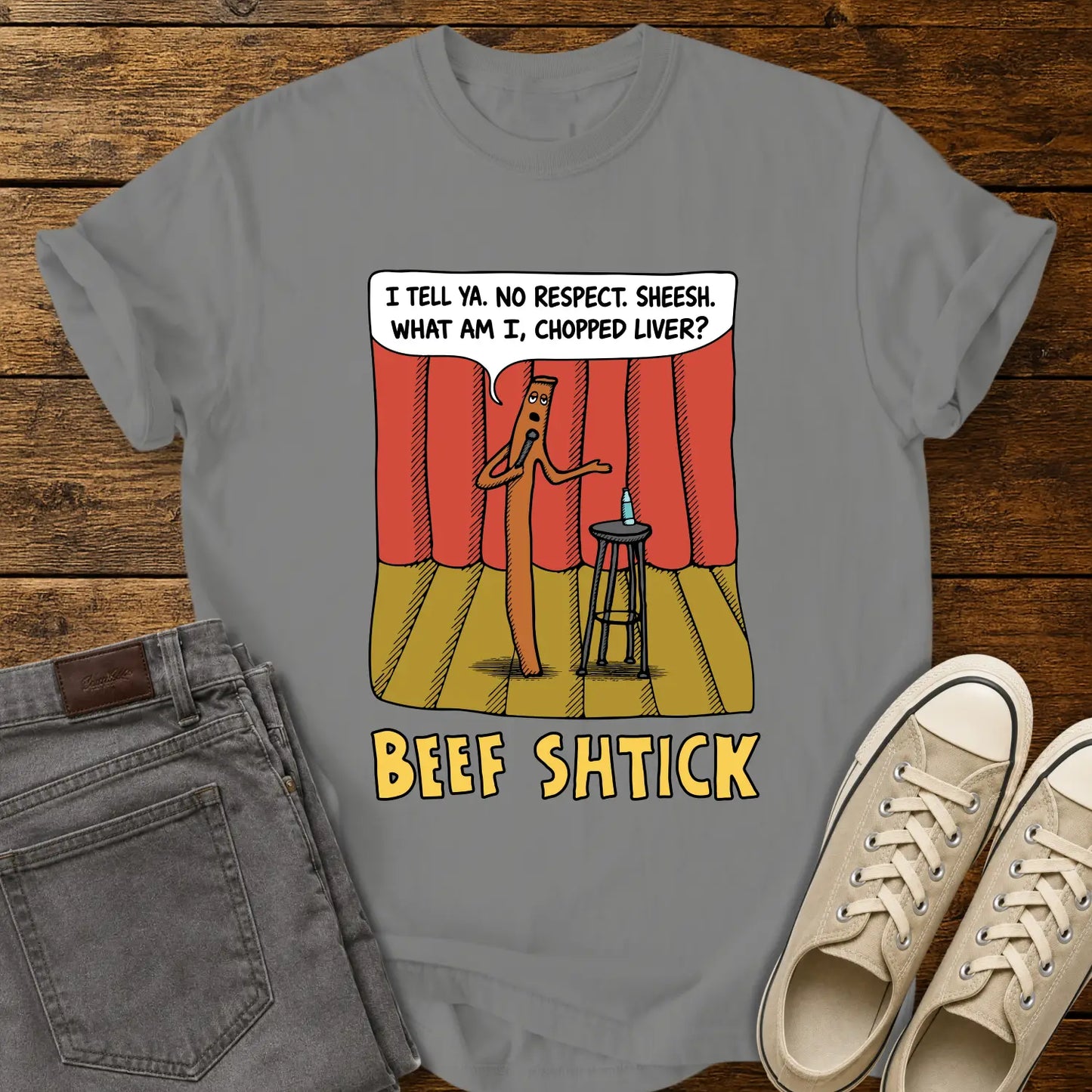 granite T-shirt featuring a humorous cartoon of a beef stick performing stand-up comedy on a stage with red curtains, a stool, microphone, and water bottle. A speech bubble reads, “I tell ya. No respect. Sheesh. What am I, chopped liver?” The design is titled “BEEF SHTICK” in bold yellow lettering. The shirt is displayed on a rustic wood plank background with folded gray jeans and cream canvas sneakers.