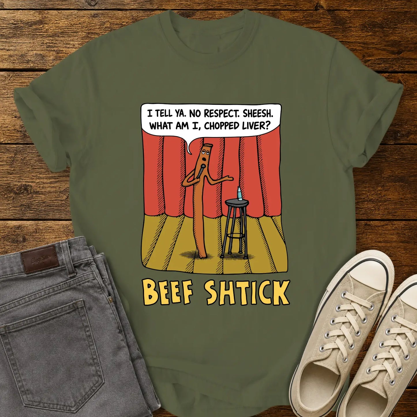 hemp green T-shirt featuring a humorous cartoon of a beef stick performing stand-up comedy on a stage with red curtains, a stool, microphone, and water bottle. A speech bubble reads, “I tell ya. No respect. Sheesh. What am I, chopped liver?” The design is titled “BEEF SHTICK” in bold yellow lettering. The shirt is displayed on a rustic wood plank background with folded gray jeans and cream canvas sneakers.
