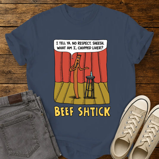 Midnight blue T-shirt featuring a humorous cartoon of a beef stick performing stand-up comedy on a stage with red curtains, a stool, microphone, and water bottle. A speech bubble reads, “I tell ya. No respect. Sheesh. What am I, chopped liver?” The design is titled “BEEF SHTICK” in bold yellow lettering. The shirt is displayed on a rustic wood plank background with folded gray jeans and cream canvas sneakers.