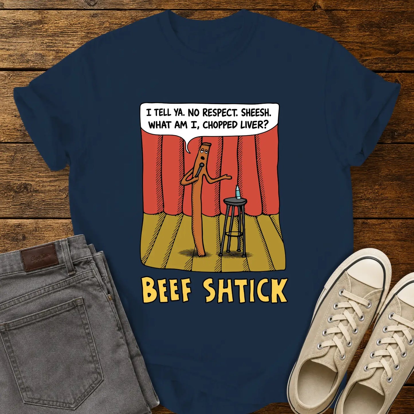 true navy blue T-shirt featuring a humorous cartoon of a beef stick performing stand-up comedy on a stage with red curtains, a stool, microphone, and water bottle. A speech bubble reads, “I tell ya. No respect. Sheesh. What am I, chopped liver?” The design is titled “BEEF SHTICK” in bold yellow lettering. The shirt is displayed on a rustic wood plank background with folded gray jeans and cream canvas sneakers.