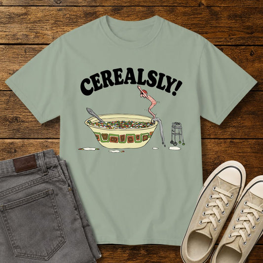 Bay green graphic T-shirt mockup featuring the phrase “Cerealsly!” above a humorous illustration of an elderly person diving into a giant bowl of cereal, styled with gray jeans and beige sneakers on a rustic wood background.