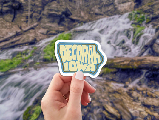 decorah iowa travel luggage sticker, hand display, mountain stream background