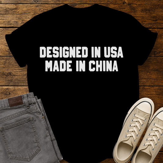 Black graphic T-shirt featuring bold white block lettering that reads “Designed in USA Made in China,” displayed on a rustic wooden background with gray jeans and beige sneakers.