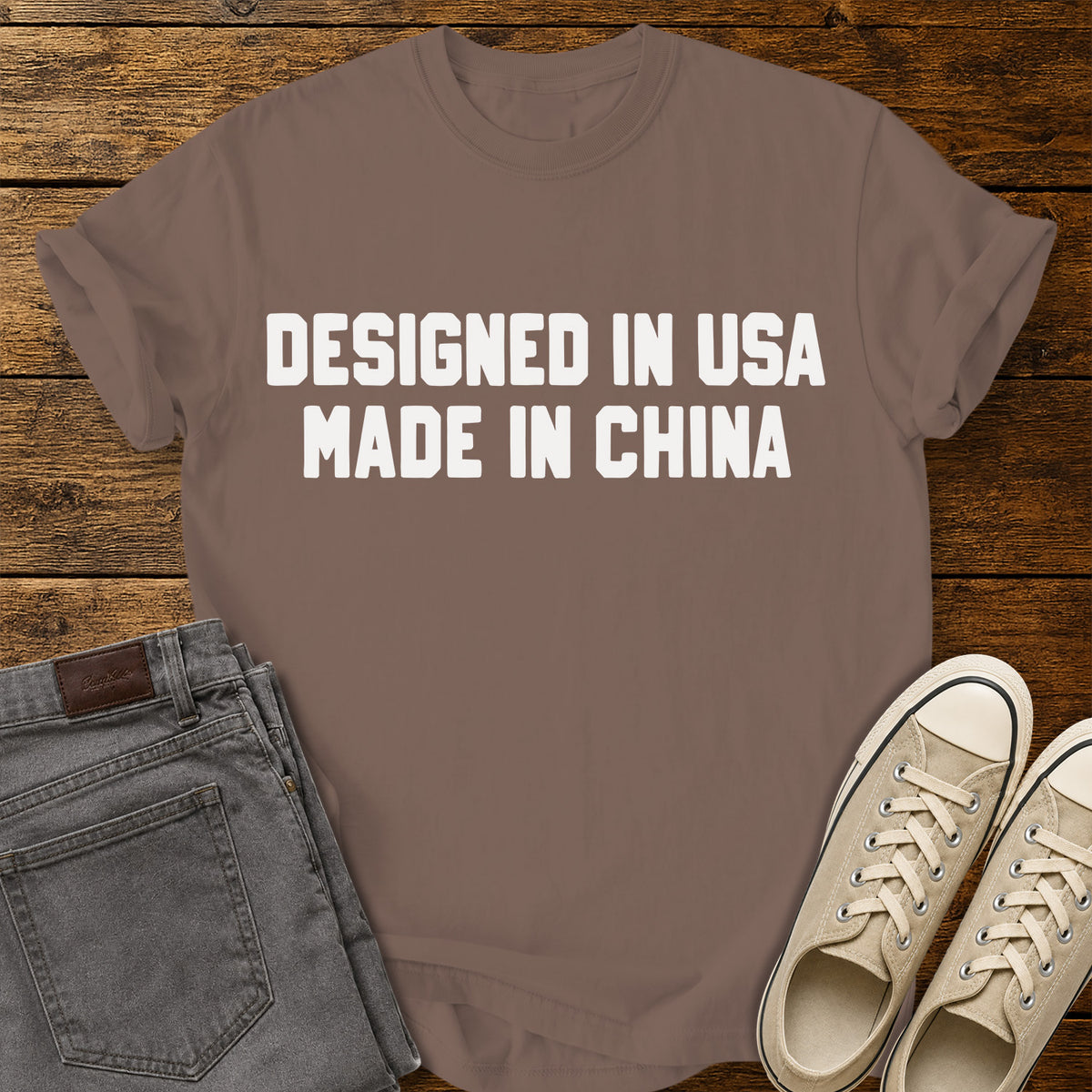 espresso brown graphic T-shirt featuring bold white block lettering that reads “Designed in USA Made in China,” displayed on a rustic wooden background with gray jeans and beige sneakers.