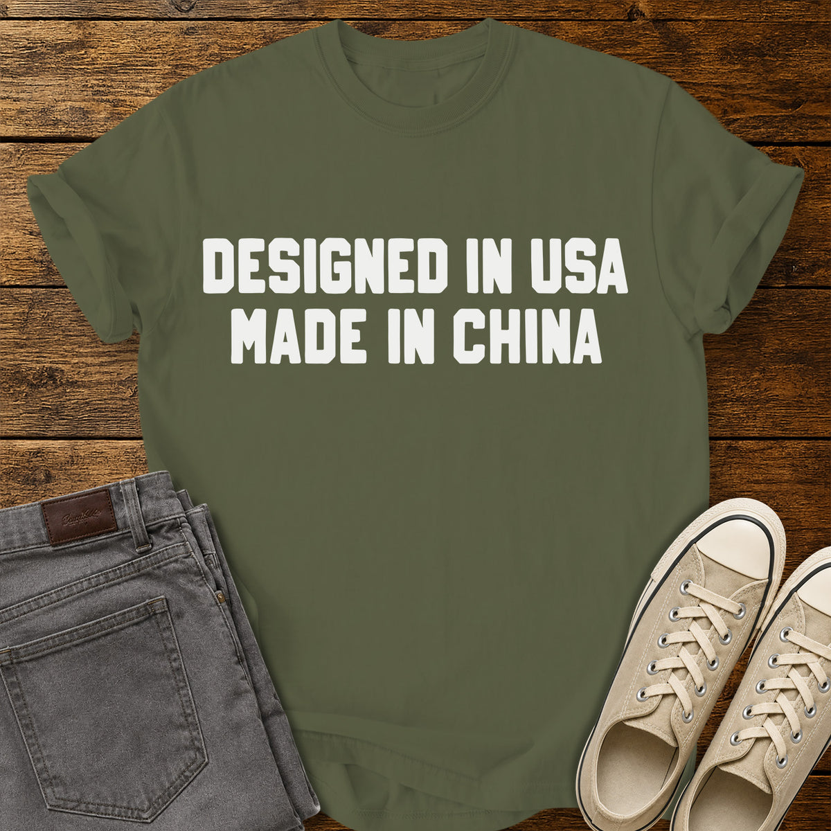 hemp green graphic T-shirt featuring bold white block lettering that reads “Designed in USA Made in China,” displayed on a rustic wooden background with gray jeans and beige sneakers.