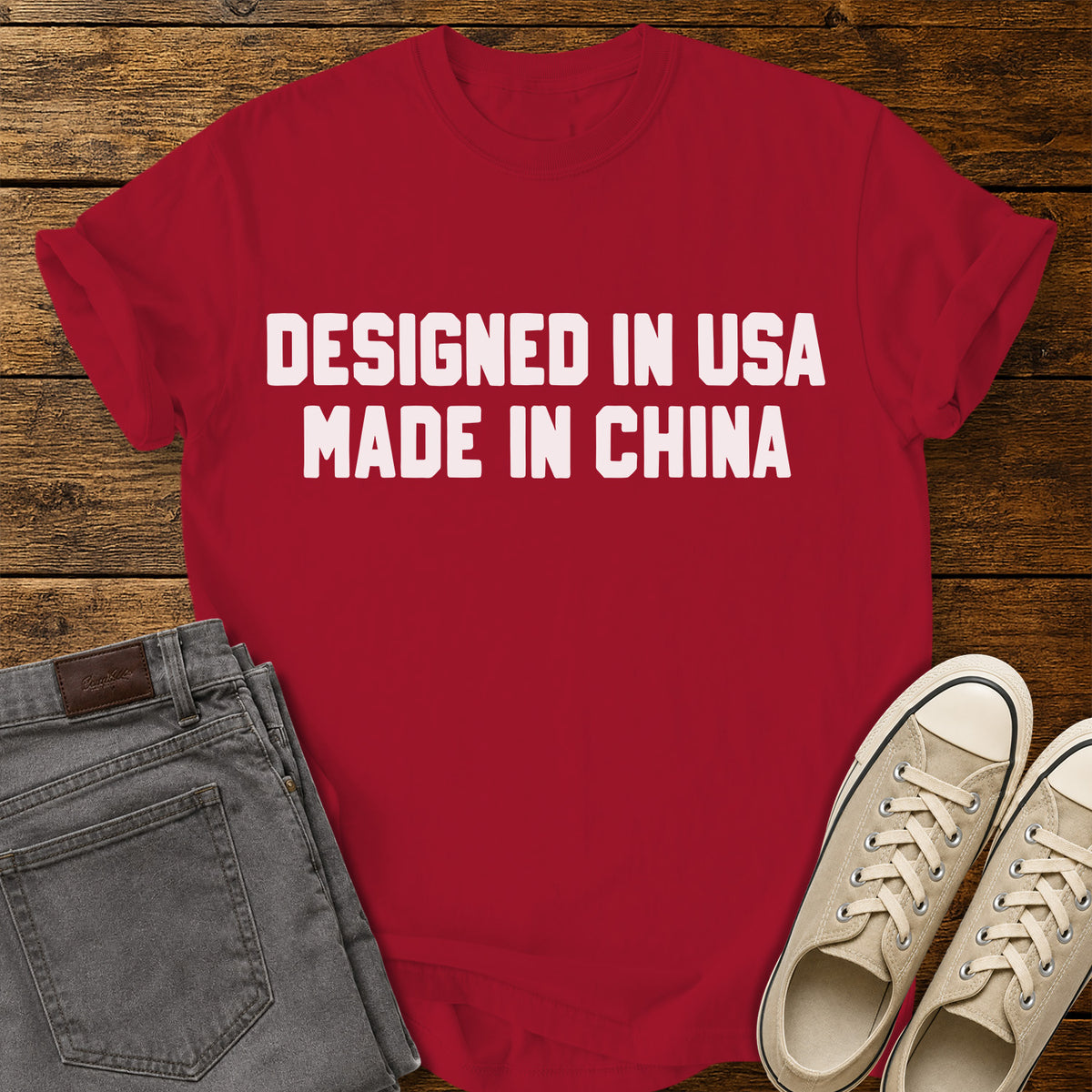 red graphic T-shirt featuring bold white block lettering that reads “Designed in USA Made in China,” displayed on a rustic wooden background with gray jeans and beige sneakers.