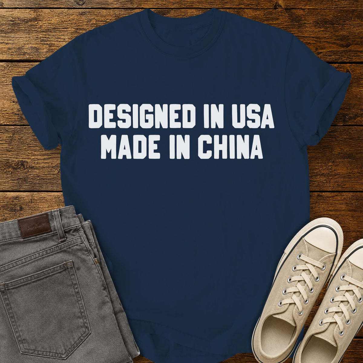 true navy blue graphic T-shirt featuring bold white block lettering that reads “Designed in USA Made in China,” displayed on a rustic wooden background with gray jeans and beige sneakers.
