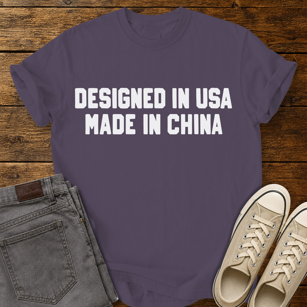 wine purple graphic T-shirt featuring bold white block lettering that reads “Designed in USA Made in China,” displayed on a rustic wooden background with gray jeans and beige sneakers. 