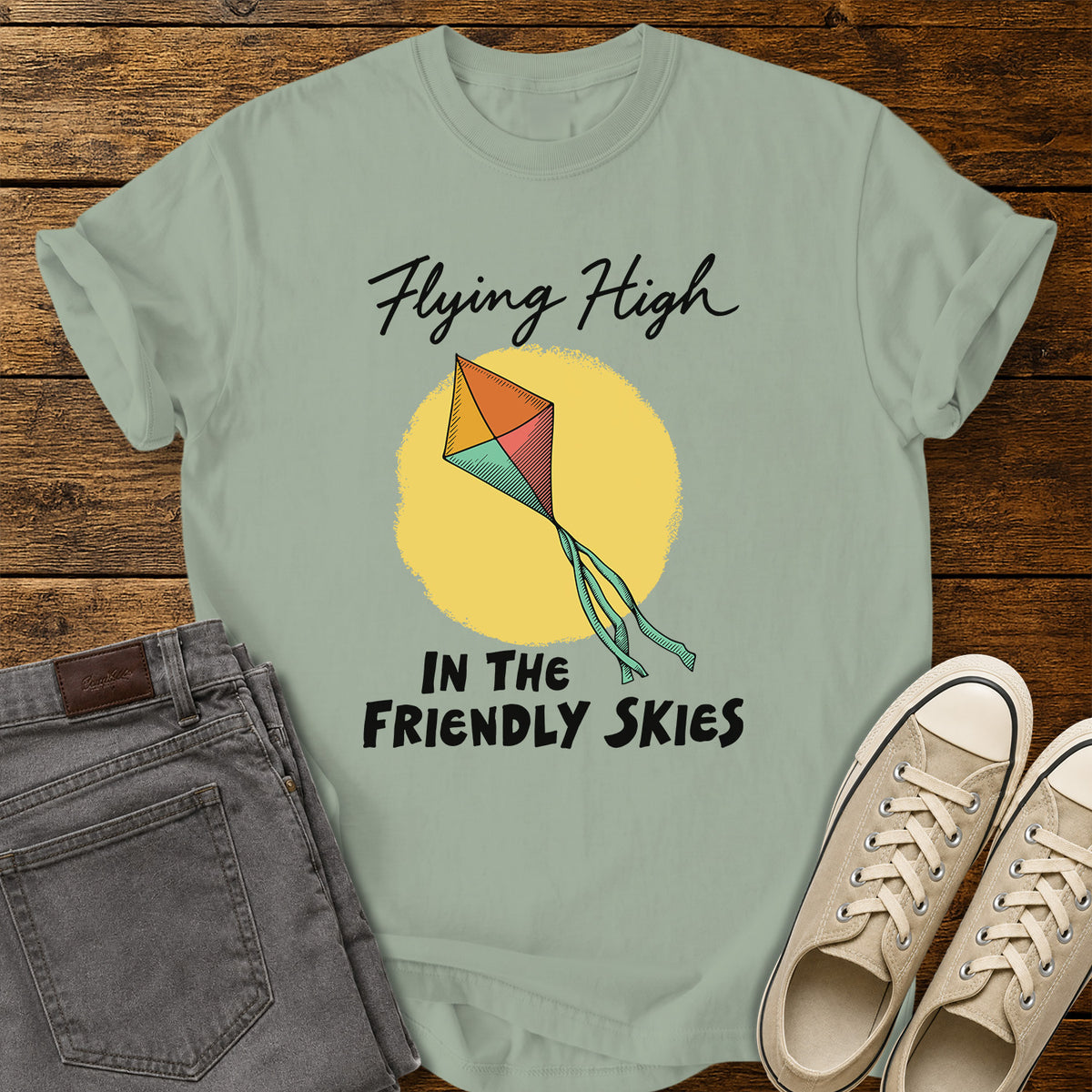 bay green T-shirt featuring a colorful retro kite flying against a yellow sun, with black script and bold lettering that reads “Flying High in the Friendly Skies,” displayed on a rustic wooden background with gray jeans and beige sneakers.
