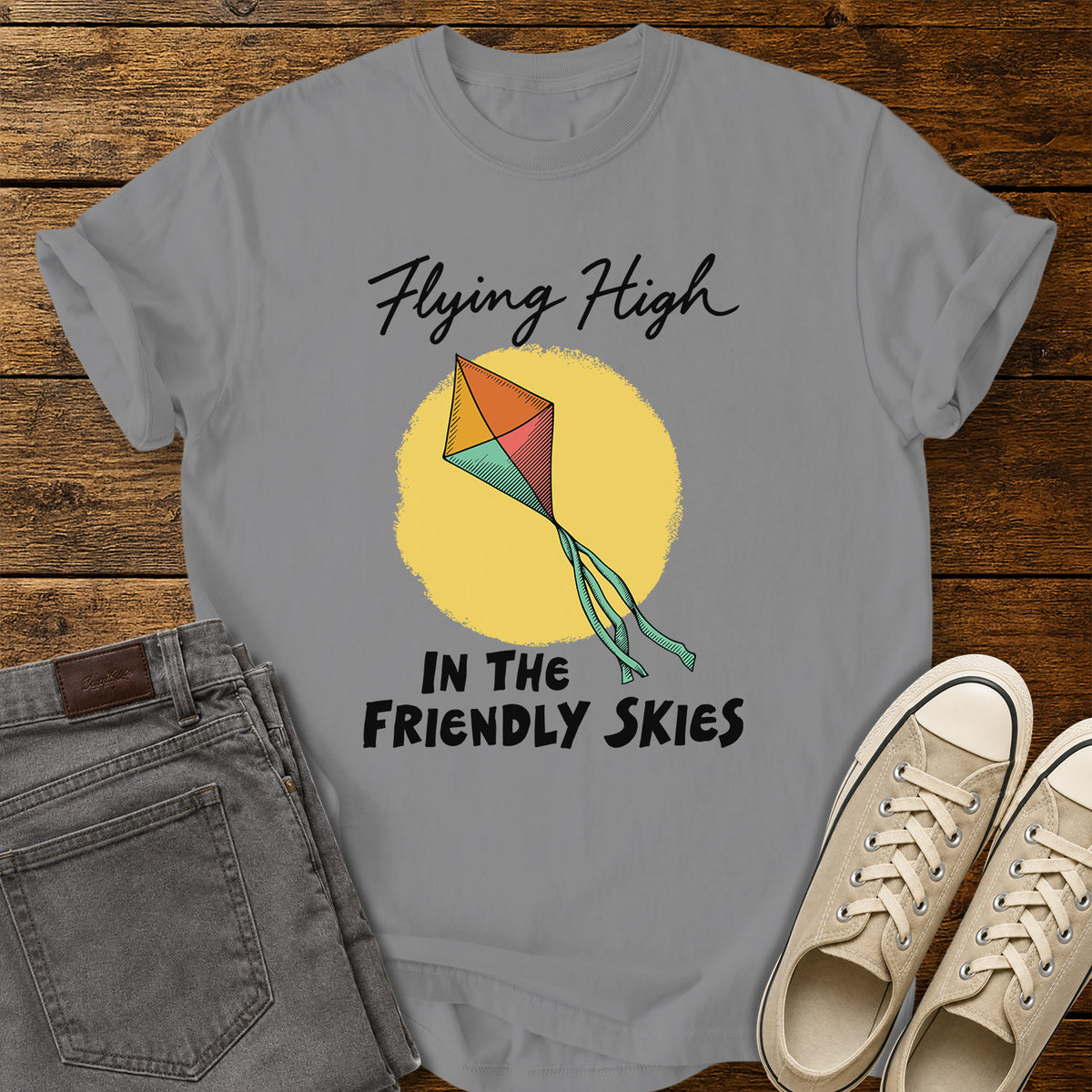 grey T-shirt featuring a colorful retro kite flying against a yellow sun, with black script and bold lettering that reads “Flying High in the Friendly Skies,” displayed on a rustic wooden background with gray jeans and beige sneakers.