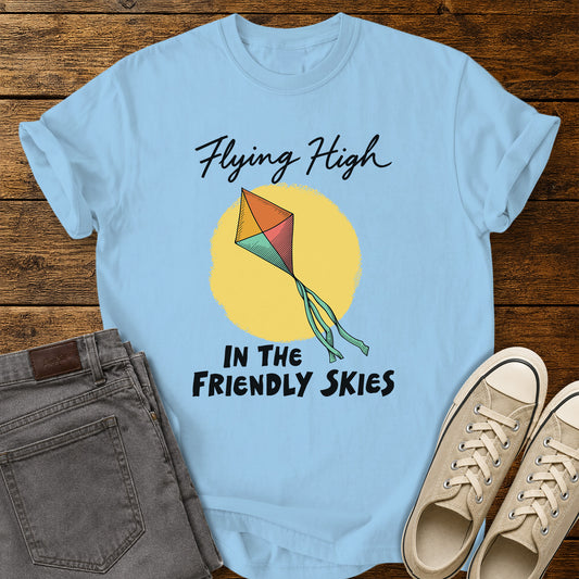 Hydrangea blue T-shirt featuring a colorful retro kite flying against a yellow sun, with black script and bold lettering that reads “Flying High in the Friendly Skies,” displayed on a rustic wooden background with gray jeans and beige sneakers.