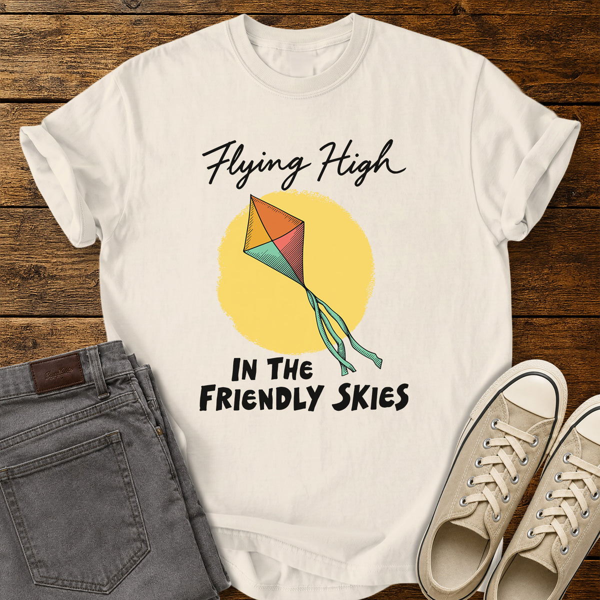 ivory T-shirt featuring a colorful retro kite flying against a yellow sun, with black script and bold lettering that reads “Flying High in the Friendly Skies,” displayed on a rustic wooden background with gray jeans and beige sneakers.
